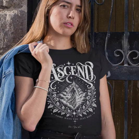 A woman holds a denim jacket over her shoulder in front of a dark wooden doorway, wearing the Ascend T-Shirt. Ascend T-Shirt is a Gildan 64000 and the color is Black.