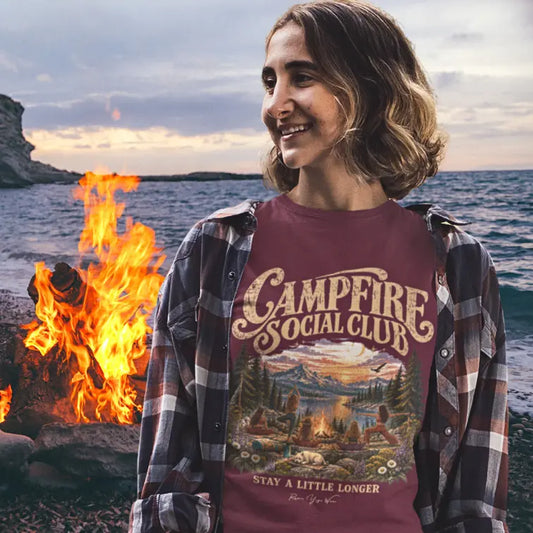 A smiling woman with a wavy bob wearing the Campfire T-Shirt under an open plaid flannel shirt, standing by a lakeside campfire at sunset, with the lakeside yoga group design on the front. Color is Heather Cardinal.