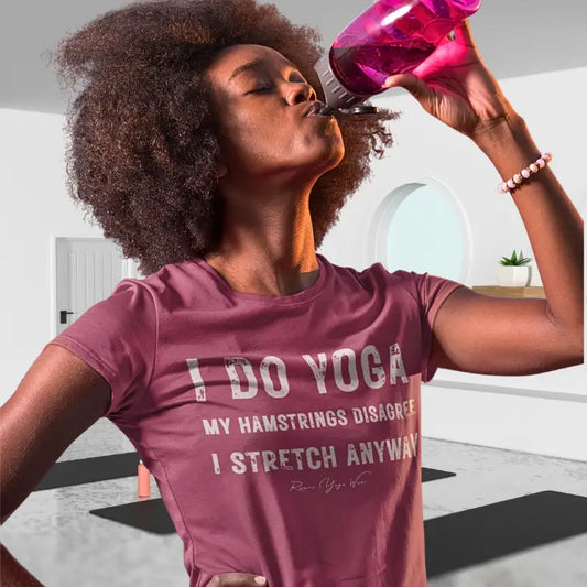 A woman with curly dark hair drinking from a pink water bottle in a bright yoga studio, wearing the Hamstrings T-Shirt with the bold stacked typography on the front. Color is Heather Raspberry.