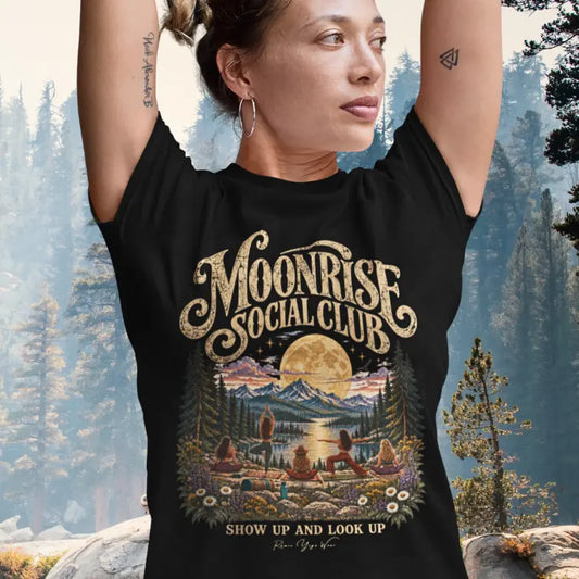A woman with her hair up and her arms raised overhead in Tree Pose wearing the Moonrise T-Shirt, standing in a sunlit pine forest, with the moonlit lake and yoga group design on the front. Color is Black.