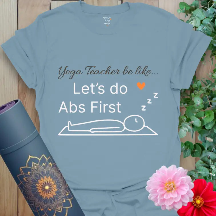 Stone Blue Abs First T-Shirt with witty yoga saying “Yoga Teacher be like… Let’s do Abs First.” Features clean white text, a minimalist stick figure in Savasana, and a signature orange heart symbol.