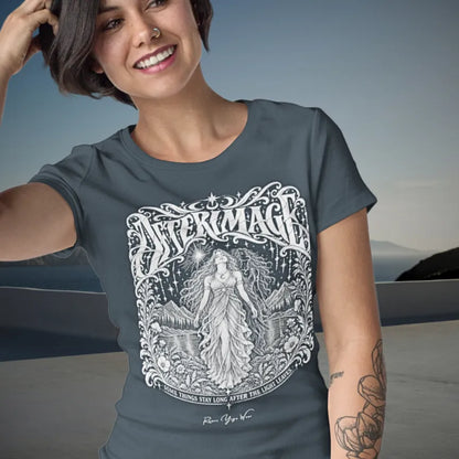 A smiling person with short dark hair and a tattooed arm, one hand raised to their hair, wearing the Afterimage T-Shirt on a coastal terrace at dusk. The tee shows ornate white linework of a figure beneath a starry sky. Color is Heather Navy.