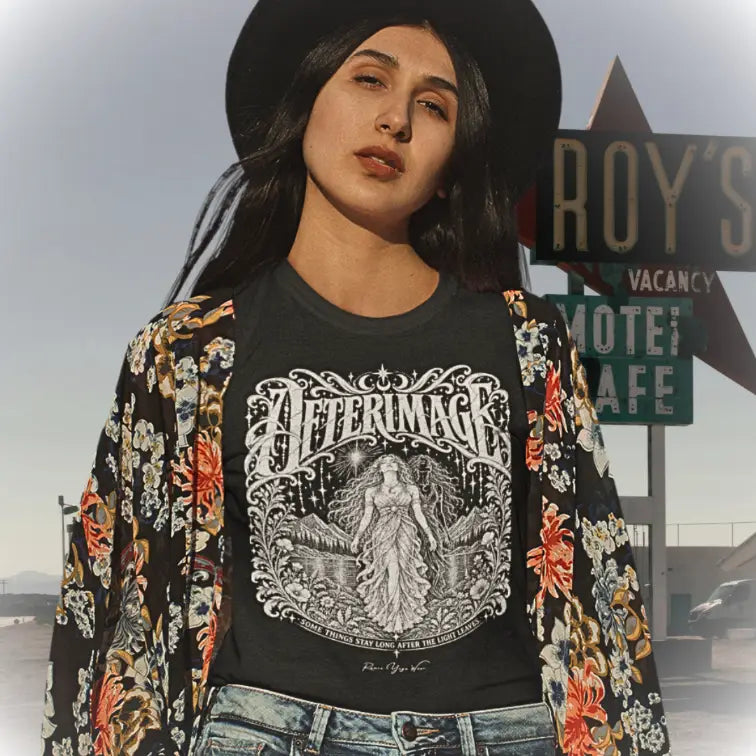 A woman in the Californian Desert wearing ai a wide-brim black hat and an open floral kimono jacket over the Afterimage T-Shirt, standing in front of a retro roadside motel sign. The tee shows ornate white linework of a figure beneath a starry sky. Tee called Afterimage T-Shirt and color is black.