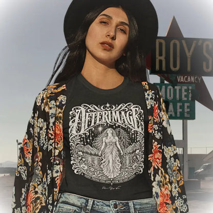 A woman in the Californian Desert wearing ai a wide-brim black hat and an open floral kimono jacket over the Afterimage T-Shirt, standing in front of a retro roadside motel sign. The tee shows ornate white linework of a figure beneath a starry sky. Tee called Afterimage T-Shirt and color is black.