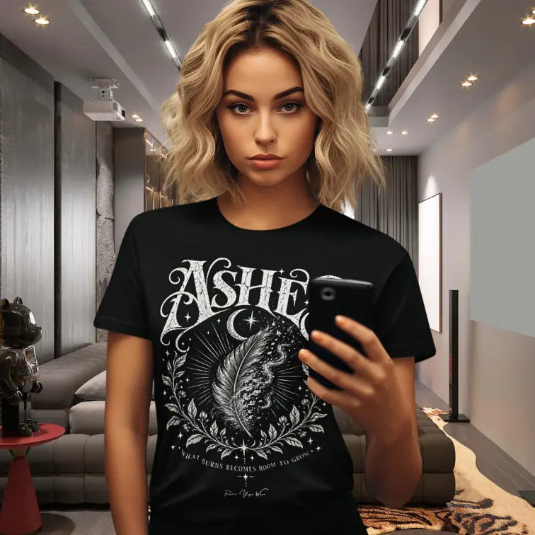 A woman with wavy blonde hair takes a mirror selfie in a modern loft-style living room, wearing the Ashes T-Shirt. Ashes T-Shirt is a Gildan 64000 and the color is Black.