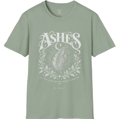 A single feather transforming into ash and ember against a dark starlit background, botanical framing below. Ashes T-Shirt flat lay is a Gildan 64000 and the color is Sage.