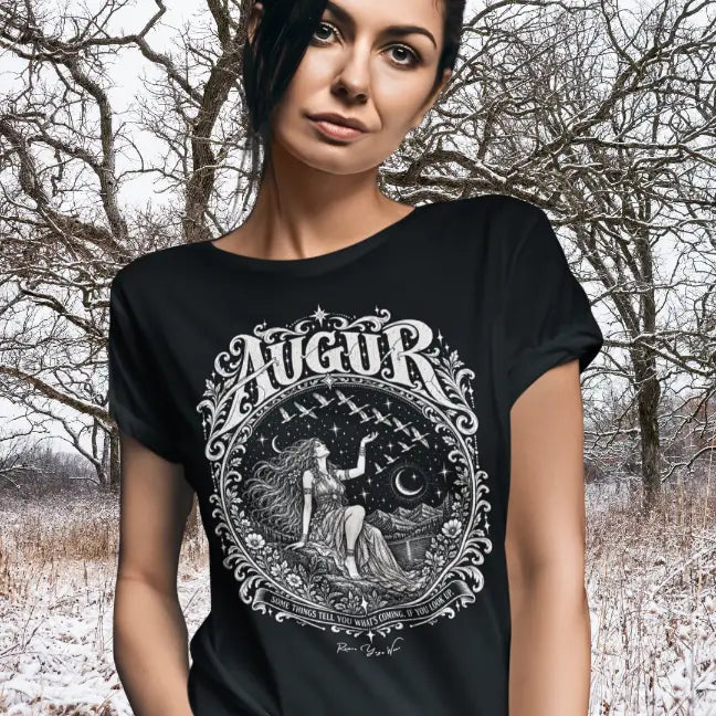 A person with long dark hair pulled back and a steady gaze, sleeves rolled, wearing the Augur T-Shirt with a white engraved seated figure and flying birds design, standing before bare winter trees and snow-covered ground. Color is Black.
