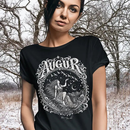 A person with long dark hair pulled back and a steady gaze, sleeves rolled, wearing the Augur T-Shirt with a white engraved seated figure and flying birds design, standing before bare winter trees and snow-covered ground. Color is Black.