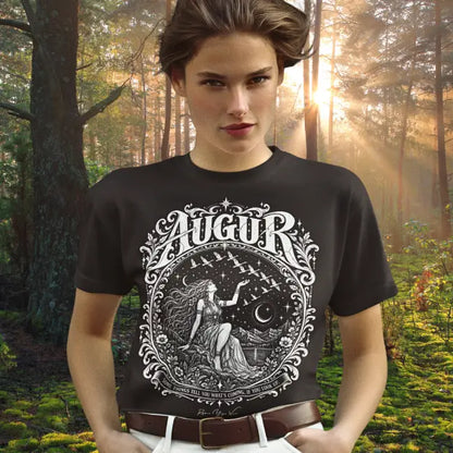 A person with swept-back brown hair and a direct gaze, hands in pockets, wearing the Augur T-Shirt with a white engraved seated figure and flying birds design, tucked into white jeans with a brown leather belt, standing in a misty pine forest with sunlight streaming through the trees. Color is Dark Chocolate.