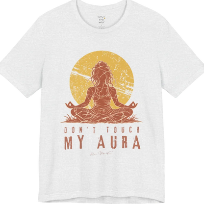 A short sleeved teed called the Aura T-Shirt, flat lay and a Bella + Canvas 3001 shirt. Simply says, Don't Touch My Aura. Design highlights a person meditating with a large bright sun in the background, all in a rustic, distressed illustration. Color is Ash.