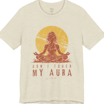 A short sleeved teed called the Aura T-Shirt, flat lay and a Bella + Canvas 3001 shirt. Simply says, Don't Touch My Aura. Design highlights a person meditating with a large bright sun in the background, all in a rustic, distressed illustration. Color is Heather Prism Natural.