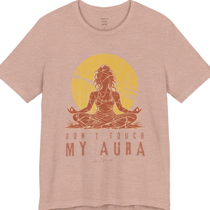 A short sleeved teed called the Aura T-Shirt, flat lay and a Bella + Canvas 3001 shirt. Simply says, Don't Touch My Aura. Design highlights a person meditating with a large bright sun in the background, all in a rustic, distressed illustration. Color is Heather Prism Peach.