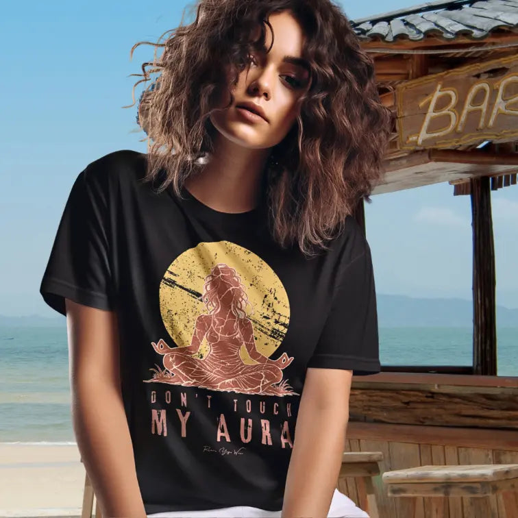 A short sleeved teed called the Aura T-Shirt, as a Bella + Canvas 3001 shirt. Simply says, Don't Touch My Aura. Design highlights a person meditating with a large bright sun in the background, all in a rustic, distressed illustration. Model is wearing her tee by the beach, relaxing.  Color is Black.
