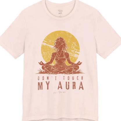 A short sleeved teed called the Aura T-Shirt, flat lay and a Bella + Canvas 3001 shirt. Simply says, Don't Touch My Aura. Design highlights a person meditating with a large bright sun in the background, all in a rustic, distressed illustration. Color is Soft Pink.