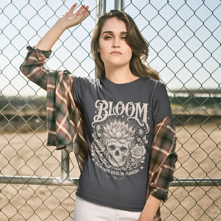 Woman wearing Bloom T-Shirt (short sleeved) outdoors against a chain-link fence, hand raised to her hair, shirt in Dark Heather Grey.