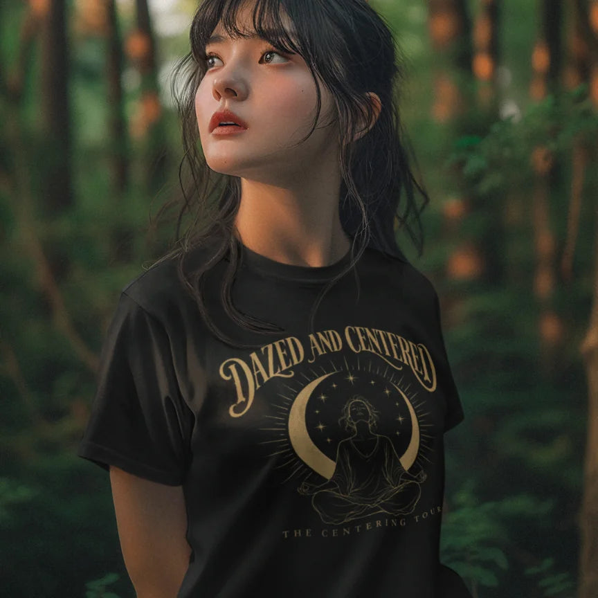 A person with long dark hair and bangs standing in a sunlit forest, wearing the Centered T-Shirt in black with the Dazed and Centered design across the chest.