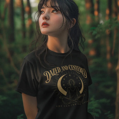 A person with long dark hair and bangs standing in a sunlit forest, wearing the Centered T-Shirt in black with the Dazed and Centered design across the chest.