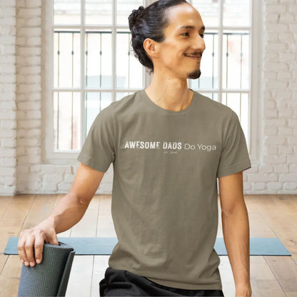 A smiling person with dark hair tied back in a bun, wearing an olive green Dads T-Shirt that reads Awesome Dads Do Yoga, sitting beside a rolled yoga mat in a bright brick-walled studio.