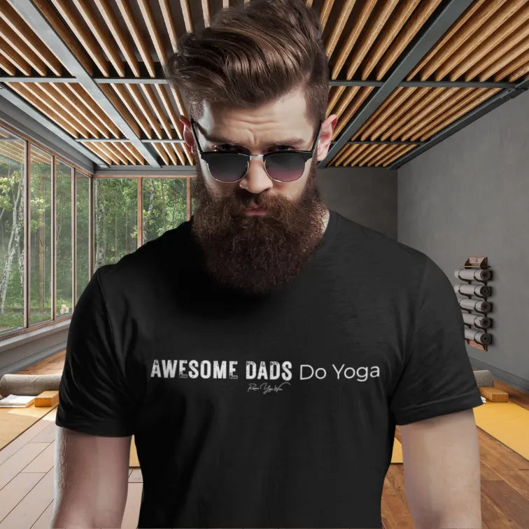 A bearded person in black sunglasses wearing a black Dads T-Shirt that reads Awesome Dads Do Yoga, standing in a sunlit yoga studio with a wooden slatted ceiling.
