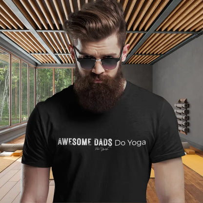 A bearded person in black sunglasses wearing a black Dads T-Shirt that reads Awesome Dads Do Yoga, standing in a sunlit yoga studio with a wooden slatted ceiling.
