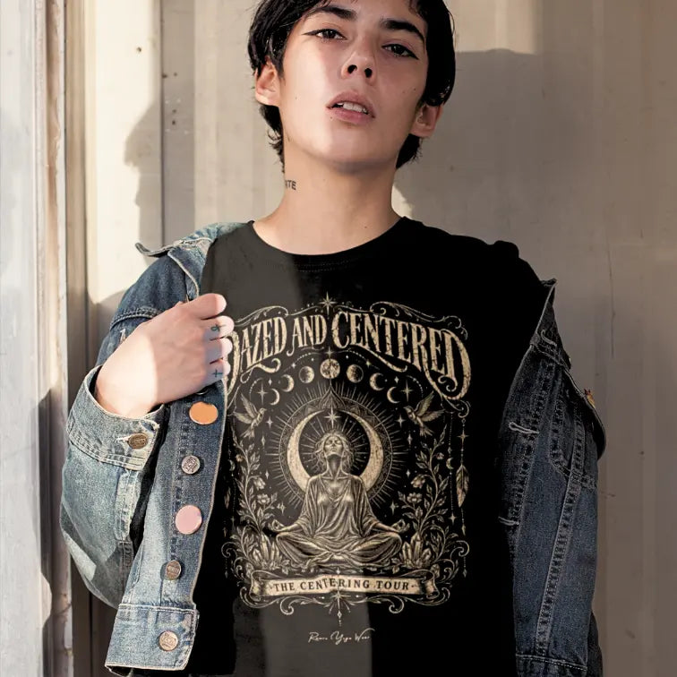 A person with dark hair standing against a pale wall in an open denim jacket, wearing the Dazed T-Shirt in black with the Dazed and Centered design across the chest.