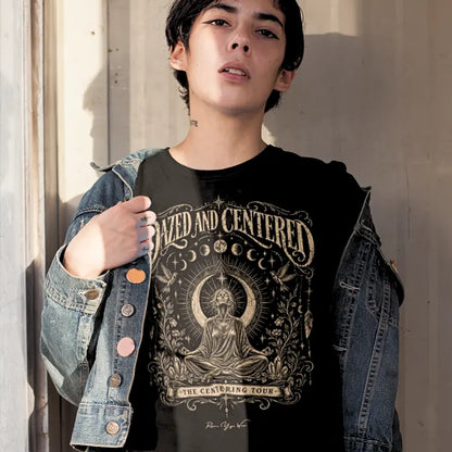 A person with dark hair standing against a pale wall in an open denim jacket, wearing the Dazed T-Shirt in black with the Dazed and Centered design across the chest.