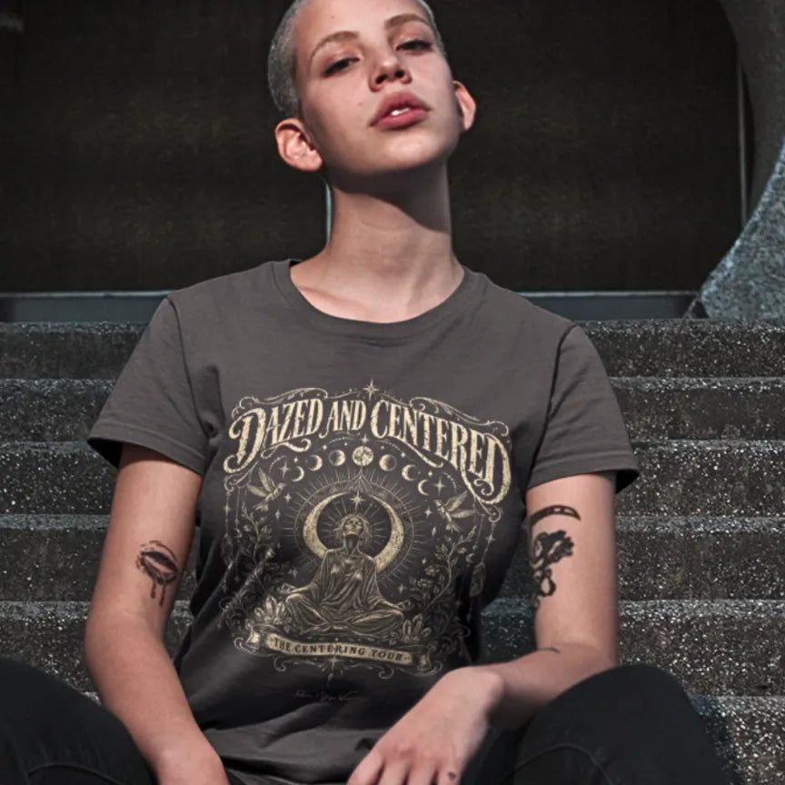 A person with a shaved head sitting on concrete steps, wearing the Dazed T-Shirt in dark chocolate with the Dazed and Centered design across the chest.