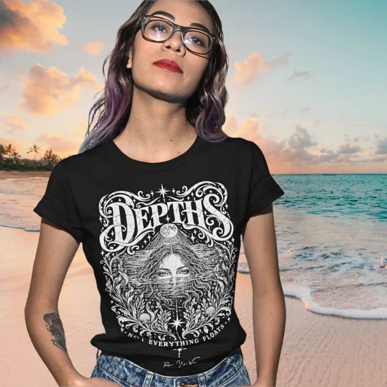 A woman in sunglasses stands on a beach at sunset with the ocean behind her, wearing the Depths T-Shirt. Depths T-Shirt is a Gildan 64000 and the color is Black.