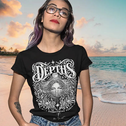 A woman in sunglasses stands on a beach at sunset with the ocean behind her, wearing the Depths T-Shirt. Depths T-Shirt is a Gildan 64000 and the color is Black.