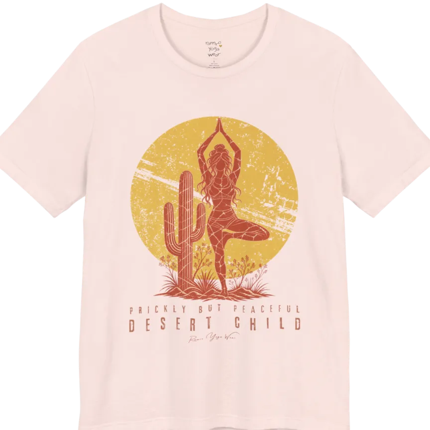 A short sleeved tee called the Desert Child T-Shirt, flat lay and a Bella + Canvas 3001 shirt. Simply says, Prickly But Peaceful, Desert Child. Design highlights a person in Tree Pose beside a desert cactus, with a large distressed sun in the background, in a rustic, distressed illustration. Color is Soft Pink.