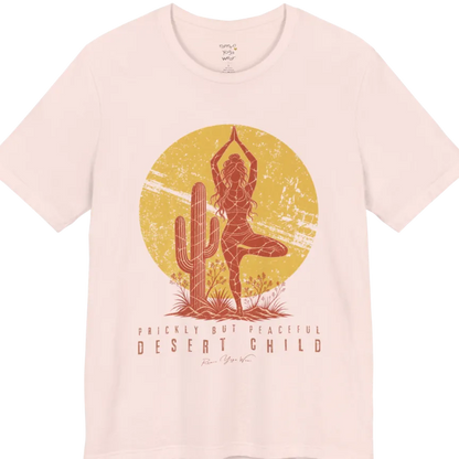 A short sleeved tee called the Desert Child T-Shirt, flat lay and a Bella + Canvas 3001 shirt. Simply says, Prickly But Peaceful, Desert Child. Design highlights a person in Tree Pose beside a desert cactus, with a large distressed sun in the background, in a rustic, distressed illustration. Color is Soft Pink.