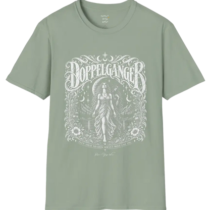 A short sleeved tee called the Doppelganger T-Shirt, flat lay and a Gildan 64000 shirt. Simply says, Even Your Shadow Doesn't Move Like You. Design highlights a woman standing before a crescent moon with a second shadow-self figure behind her, framed by mountains, florals, and ornate engraved linework border. Color is Sage.