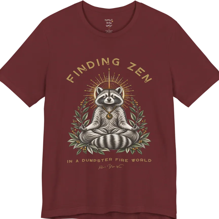 A short sleeved Tee of a meditating raccoon illustration with a gold sunburst halo and botanical leaf framing. Dumpster T-Shirt flat lay is a Bella+Canvas 3001 and the color is Heather Cardinal.