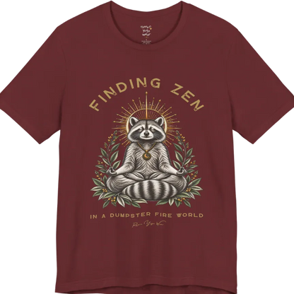 A short sleeved Tee of a meditating raccoon illustration with a gold sunburst halo and botanical leaf framing. Dumpster T-Shirt flat lay is a Bella+Canvas 3001 and the color is Heather Cardinal.