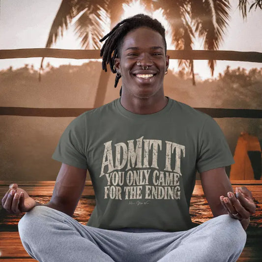 A smiling man with dreadlocks sitting cross-legged in a meditation pose on a wooden deck at sunset, wearing the Ending T-Shirt with the stacked Admit It typography on the front. Color is Heather Military Green.