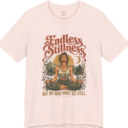 A short sleeved tee, showing an Illustration of a woman seated in lotus meditation pose in front of mountains, pine trees, and a crescent moon, framed by botanical leaves and flowers. Endless T-Shirt flat lay is a Bella+Canvas 3001 and the color is Soft Pink.