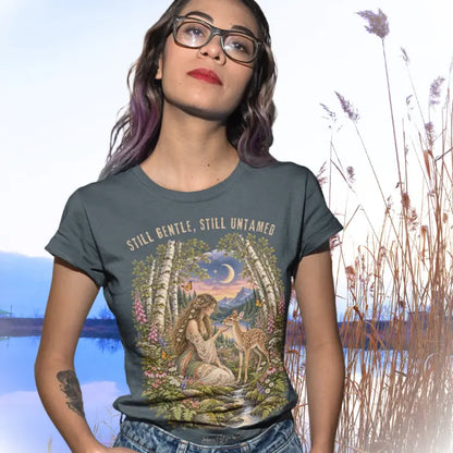 A short sleeved tee called the Gentle T-Shirt, worn on a model with glasses and long wavy hair standing beside a calm lake with tall golden reeds under a pale sky. Simply says, Still Gentle, Still Untamed. Design highlights a woman with wildflowers in her hair kneeling in a birch forest, gently cupping the chin of a spotted fawn, with foxgloves, ferns and a small stream below a crescent moon at twilight, in a full-color painted illustration style. Color is Dark Heather Grey.