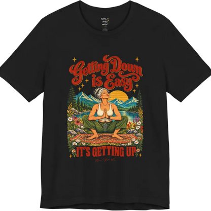 A short sleeved tee called the Getting Up T-Shirt, flat lay and a soft cotton blend shirt. Simply says, Getting Down is Easy, It's Getting Up. Design highlights retro red script lettering above a silver-haired person in a deep yoga squat with hands in prayer on a patterned mat, with a sun, mountains, a lake and wildflowers, bold red lettering for IT'S GETTING UP and a small script signature. Color is Black.
