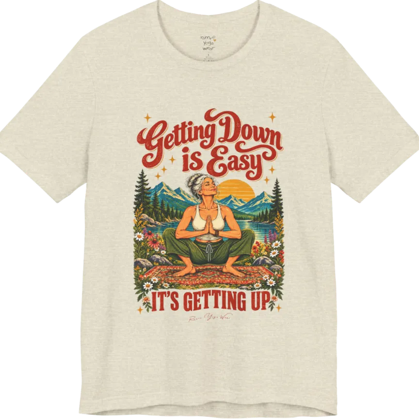 A short sleeved tee called the Getting Up T-Shirt, flat lay and a soft cotton blend shirt. Simply says, Getting Down is Easy, It's Getting Up. Design highlights retro red script lettering above a silver-haired person in a deep yoga squat with hands in prayer on a patterned mat, with a sun, mountains, a lake and wildflowers, bold red lettering for IT'S GETTING UP and a small script signature. Color is Heather Prism Natural.
