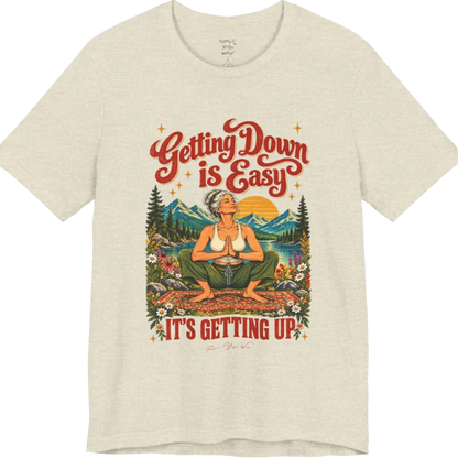 A short sleeved tee called the Getting Up T-Shirt, flat lay and a soft cotton blend shirt. Simply says, Getting Down is Easy, It's Getting Up. Design highlights retro red script lettering above a silver-haired person in a deep yoga squat with hands in prayer on a patterned mat, with a sun, mountains, a lake and wildflowers, bold red lettering for IT'S GETTING UP and a small script signature. Color is Heather Prism Natural.