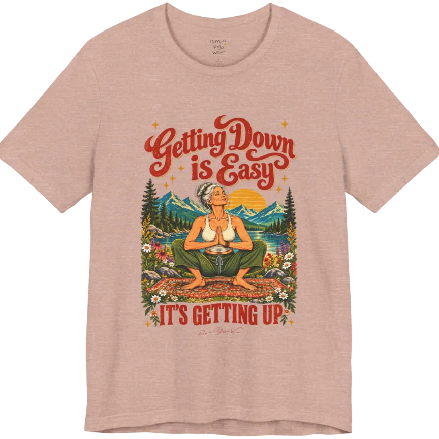 A short sleeved tee called the Getting Up T-Shirt, flat lay and a soft cotton blend shirt. Simply says, Getting Down is Easy, It's Getting Up. Design highlights retro red script lettering above a silver-haired person in a deep yoga squat with hands in prayer on a patterned mat, with a sun, mountains, a lake and wildflowers, bold red lettering for IT'S GETTING UP and a small script signature. Color is Heather Prism Peach. 