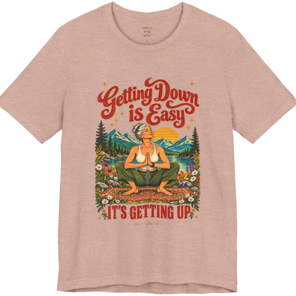 A short sleeved tee called the Getting Up T-Shirt, flat lay and a soft cotton blend shirt. Simply says, Getting Down is Easy, It's Getting Up. Design highlights retro red script lettering above a silver-haired person in a deep yoga squat with hands in prayer on a patterned mat, with a sun, mountains, a lake and wildflowers, bold red lettering for IT'S GETTING UP and a small script signature. Color is Heather Prism Peach. 