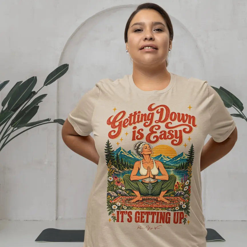 A person with dark hair standing with their hands behind their back in a bright studio with a leafy plant and an arched wall behind them, wearing a tan Getting Up T-Shirt that says Getting Down is Easy, It's Getting Up.