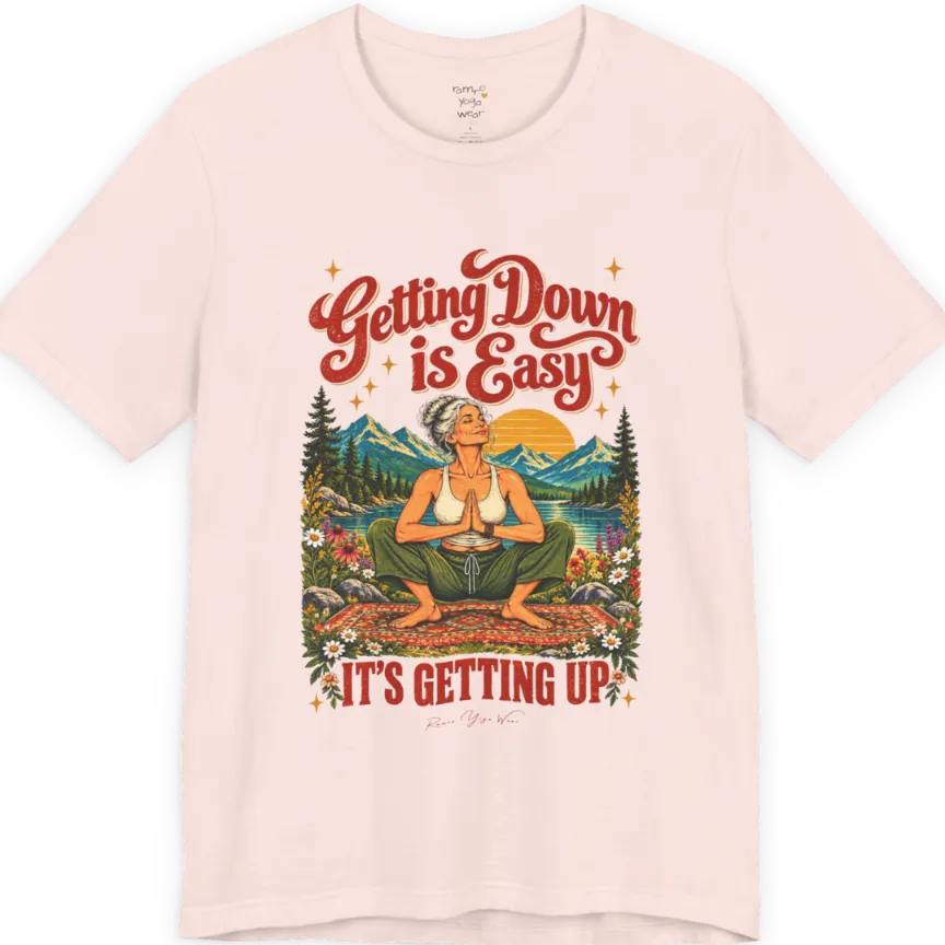 A short sleeved tee called the Getting Up T-Shirt, flat lay and a soft cotton blend shirt. Simply says, Getting Down is Easy, It's Getting Up. Design highlights retro red script lettering above a silver-haired person in a deep yoga squat with hands in prayer on a patterned mat, with a sun, mountains, a lake and wildflowers, bold red lettering for IT'S GETTING UP and a small script signature. Color is Soft Pink.