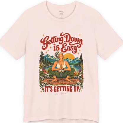 A short sleeved tee called the Getting Up T-Shirt, flat lay and a soft cotton blend shirt. Simply says, Getting Down is Easy, It's Getting Up. Design highlights retro red script lettering above a silver-haired person in a deep yoga squat with hands in prayer on a patterned mat, with a sun, mountains, a lake and wildflowers, bold red lettering for IT'S GETTING UP and a small script signature. Color is Soft Pink.
