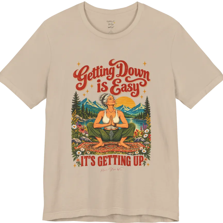 A short sleeved tee called the Getting Up T-Shirt, flat lay and a soft cotton blend shirt. Simply says, Getting Down is Easy, It's Getting Up. Design highlights retro red script lettering above a silver-haired person in a deep yoga squat with hands in prayer on a patterned mat, with a sun, mountains, a lake and wildflowers, bold red lettering for IT'S GETTING UP and a small script signature. Color is Tan. 