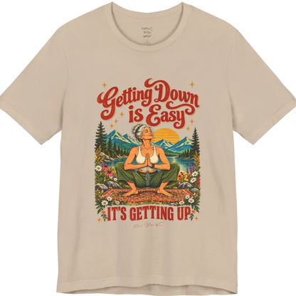 A short sleeved tee called the Getting Up T-Shirt, flat lay and a soft cotton blend shirt. Simply says, Getting Down is Easy, It's Getting Up. Design highlights retro red script lettering above a silver-haired person in a deep yoga squat with hands in prayer on a patterned mat, with a sun, mountains, a lake and wildflowers, bold red lettering for IT'S GETTING UP and a small script signature. Color is Tan. 
