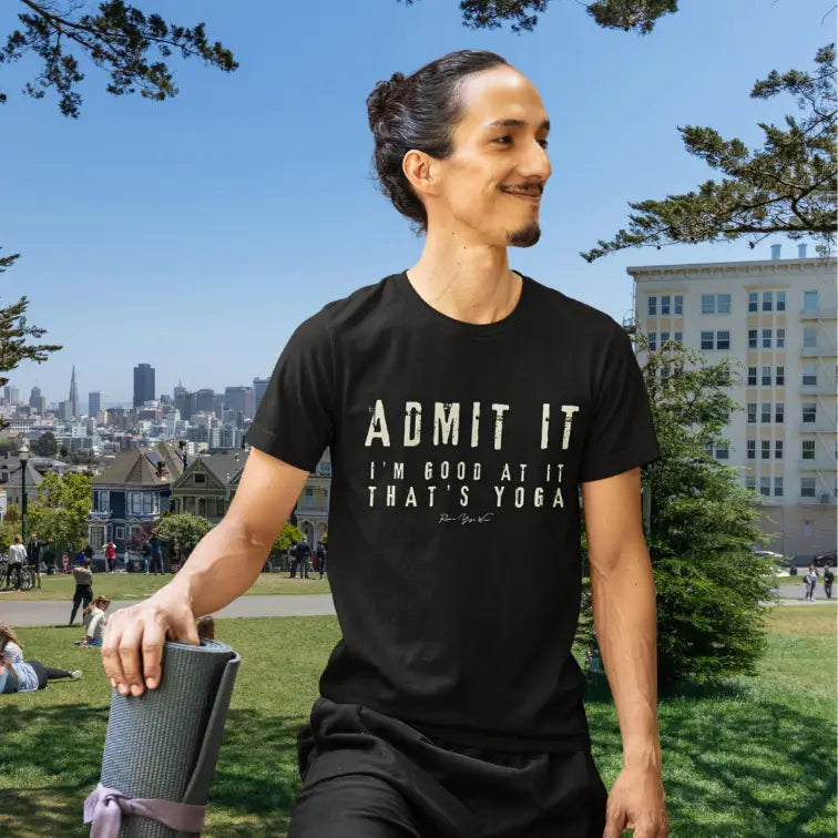 A yogi with their hair tied up in a bun, his yoga mat, and ready to work out in the park, with a city skyline behind, wearing the Good T-Shirt. The tee shows bold distressed cream lettering that says Admit It, I'm Good At It, That's Yoga. Color is black.