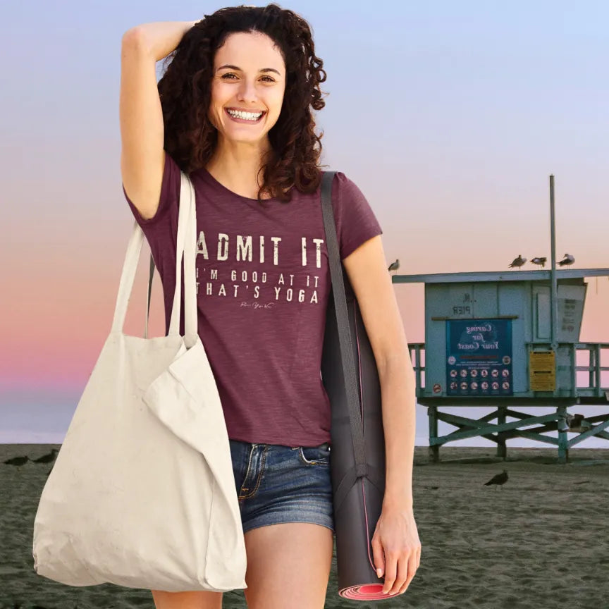 A smiling person with curly hair carrying a canvas tote bag and yoga mat on a beach at sunset with a lifeguard tower behind, wearing the Good T-Shirt. The tee shows bold distressed cream lettering that says Admit It, I'm Good At It, That's Yoga. Color is heather cardinal.