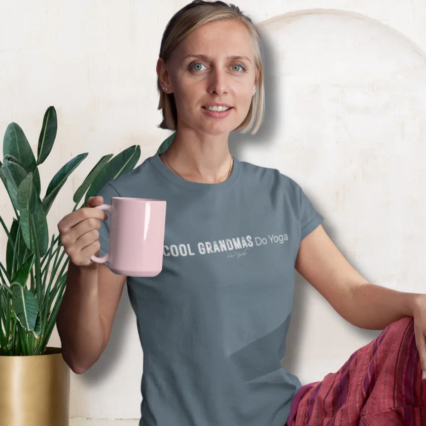 A smiling person with blonde hair holding a pink mug and sitting beside leafy plants in a bright room, wearing a the Heather Slate Grandmas T-Shirt that says Cool Grandmas Do Yoga.